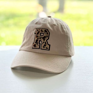 May include: A beige baseball cap with a black and brown leopard print letter "R" on the front.