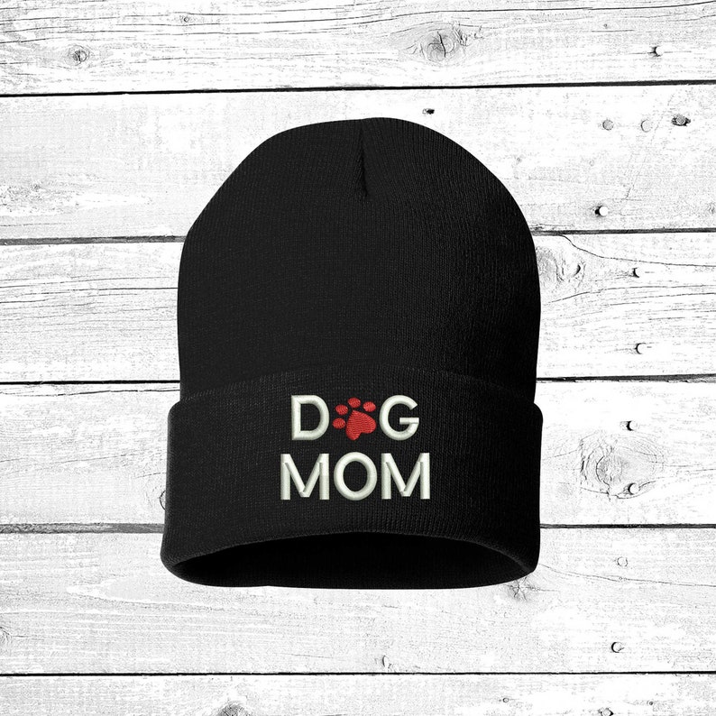 May include: A black beanie with the embroidered text "DOG MOM" in white, with a red paw print replacing the "O" in "DOG". The hat is made of a soft, knitted material.