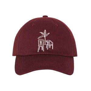 May include: A maroon baseball cap with a white embroidered design of a palm tree and two chairs.