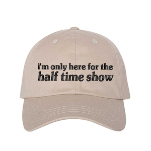 May include: A beige baseball cap with the text "I'm only here for the half time show" in black. The cap has a curved brim and a button on top.