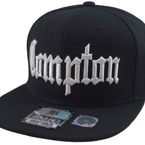 May include: Black and white snap-back baseball cap with the word "Compton" embroidered in white on the front. The cap has a black brim and a black crown.