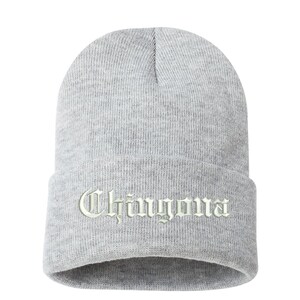May include: A light gray beanie hat with the word "Chingona" embroidered in a light green, gothic-style font. The hat is made of a knitted material and has a folded brim.