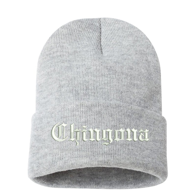 May include: A light gray beanie hat with the word "Chingona" embroidered in a light green, gothic-style font. The hat is made of a knitted material and has a folded brim.