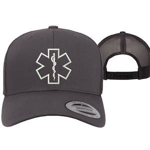 May include: A dark gray trucker hat with a white embroidered medical symbol, the Star of Life, on the front. The hat has a black mesh back and a black adjustable snap closure.