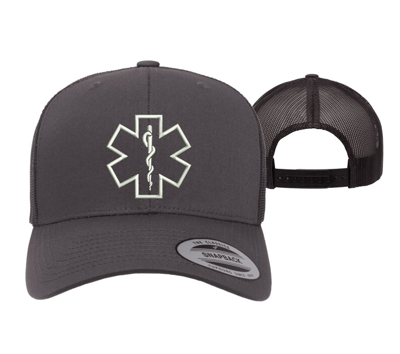May include: A dark gray trucker hat with a white embroidered medical symbol, the Star of Life, on the front. The hat has a black mesh back and a black adjustable snap closure.