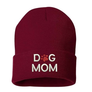 May include: A burgundy beanie with the embroidered text "DOG MOM" in white. The letter "O" in "DOG" is replaced with a red paw print. The hat is made of a soft, knitted material.