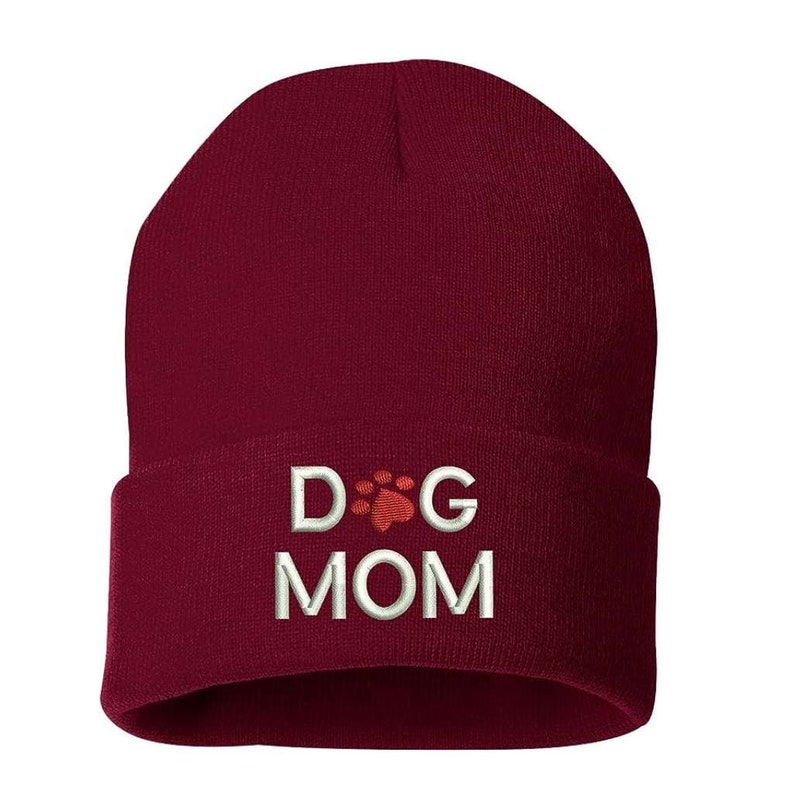 May include: A burgundy beanie with the embroidered text "DOG MOM" in white. The letter "O" in "DOG" is replaced with a red paw print. The hat is made of a soft, knitted material.