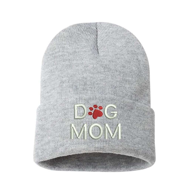 May include: A light gray beanie with the embroidered text "DOG MOM". The word "DOG" features a red paw print design in place of the letter "O". The beanie is made of a soft, knitted material.