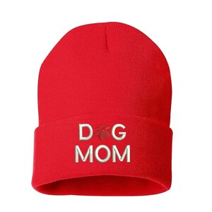 May include: A bright red beanie hat with the embroidered text "DOG MOM" in white, with a paw print graphic. The hat is a solid color and appears to be made of a soft, knitted material.