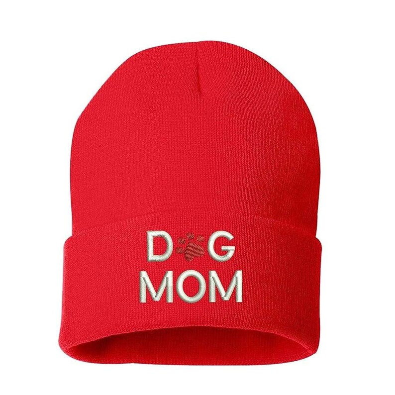 May include: A bright red beanie hat with the embroidered text "DOG MOM" in white, with a paw print graphic. The hat is a solid color and appears to be made of a soft, knitted material.