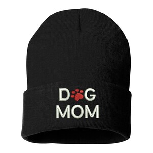 May include: Black beanie hat with the embroidered text "DOG MOM". The word "DOG" is separated by a red paw print from the word "MOM". The hat is made of a soft, knitted material.