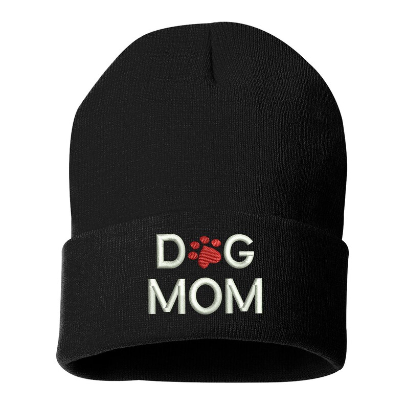 May include: Black beanie hat with the embroidered text "DOG MOM". The word "DOG" is separated by a red paw print from the word "MOM". The hat is made of a soft, knitted material.