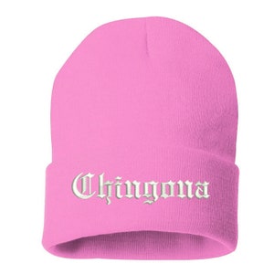 May include: A pink beanie with the word "Chingona" embroidered in white, gothic-style lettering. The hat is a solid, vibrant pink color and appears to be made of a soft, knitted material. The folded brim displays the embroidered text.