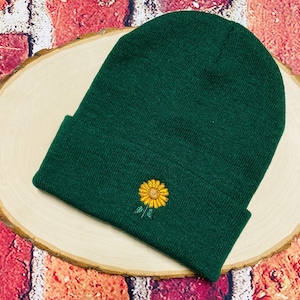 May include: A dark green beanie with a small embroidered sunflower in yellow and green.