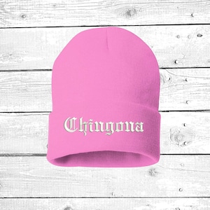 May include: A pink beanie with the word "Chingona" embroidered in white, gothic-style lettering. The hat is made of a soft, knitted material and is designed to be worn on the head. The background is a white wooden surface.