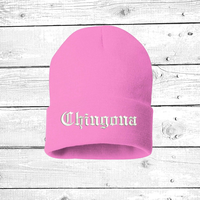 May include: A pink beanie with the word "Chingona" embroidered in white, gothic-style lettering. The hat is made of a soft, knitted material and is designed to be worn on the head. The background is a white wooden surface.