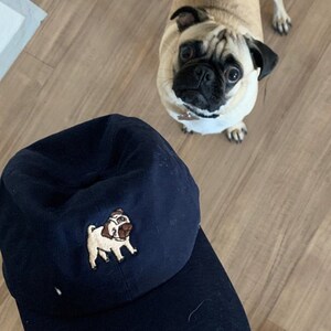 May include: A dark blue baseball cap with an embroidered image of a pug dog. The pug is light brown with a black nose and eyes.