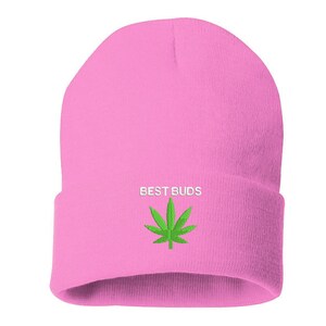 May include: A pink beanie with the embroidered text "BEST BUDS" above a green cannabis leaf. The beanie is a solid light pink color and is made of a soft, knitted material.