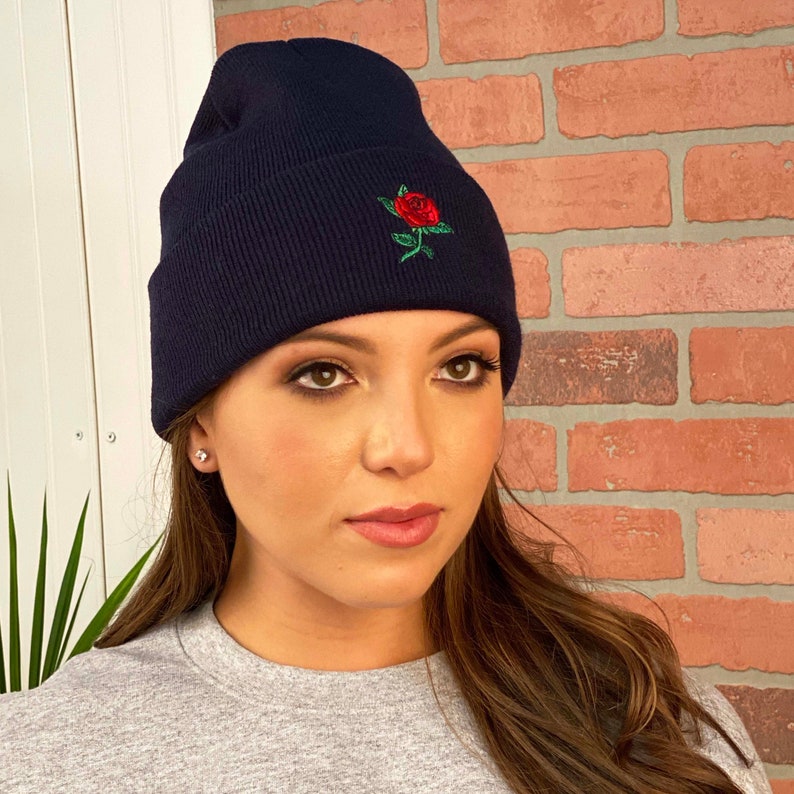 May include: Navy blue beanie with a red rose embroidered on the front. The beanie is made of a ribbed knit material. The rose has green leaves and a red flower.