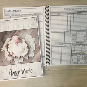 May include: A white planner with a woodgrain background and a photo of a baby girl in a basket. The planner has the text "January 2022" and "Alyssa Marie" on the cover. The planner is open to a page with a list of expenses and income.