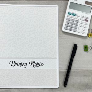 May include: A white spiral bound notebook with a leopard print design. The notebook has the name "Brinkley Marie" written on the front cover.