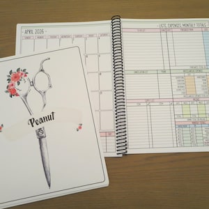 May include: A spiral-bound planner with a white cover featuring a black and white illustration of scissors adorned with pink and red flowers and the name Peanut. The open pages display an April 2026 calendar and a lists, expenses, and monthly totals page.