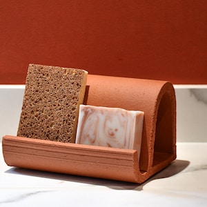 Peut inclure: Un porte-savon en terre cuite avec un design incurvé contient une éponge brune et une barre de savon blanc avec des tourbillons bruns. Le porte-savon est posé sur un comptoir en marbre blanc.