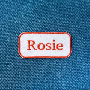 May include: A rectangular white embroidered patch with rounded corners and a bright orange border. The name Rosie is stitched in the center in a matching orange thread. The patch is displayed against a textured blue denim fabric background.