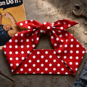 May include: A red fabric headband featuring a white polka dot pattern and a knotted bow detail at the top center. The headband is positioned horizontally, displaying a wide band design with two pointed ends tied into a bow shape.