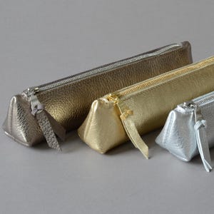 Può includere: Tre astucci metallici color oro, argento e bronzo. Ogni astuccio ha una forma triangolare e una chiusura a cerniera con linguetta abbinata. Gli astucci sono realizzati in materiale strutturato e sono disposti in fila.