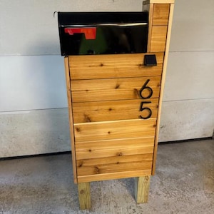 May include: A wooden mailbox stand with a black mailbox on top. The stand is made of cedar wood and has a natural finish. The mailbox has a red flag. The stand has the number "665" on the front.