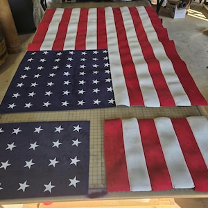 May include: A large American flag composed of red, white, and blue fabric sections laid out flat. The design features a blue canton with white stars and alternating horizontal red and white stripes, with separate fabric pieces visible below.
