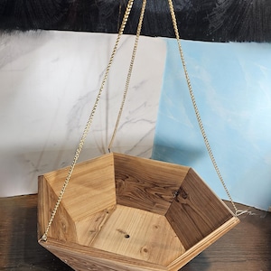 May include: A geometric wooden hanging planter with a natural finish. The planter is suspended by gold-colored chains. The design features multiple angled sides, creating a unique shape. Ideal for displaying plants or other decorative items.