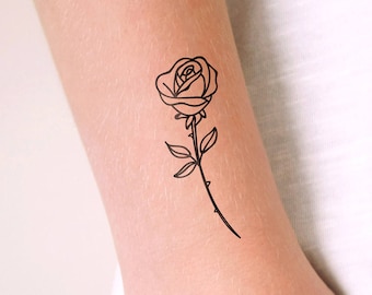 Könnte beinhalten: Schwarze Rosen-Tätowierung auf einem Arm. Die Tätowierung ist eine detaillierte Umrisszeichnung einer Rose mit Blättern, die auf dem Unterarm platziert ist. Die Person trägt ein weißes Oberteil und einen goldenen Ring.