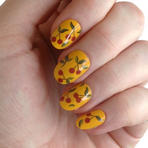 May include: A hand with yellow nail polish featuring a cherry design. The cherries are red with green stems and leaves.