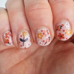 May include: Close-up of a hand with five fingernails painted in a light peach color. Each nail has a different floral design featuring white, orange, and yellow flowers with black stems.
