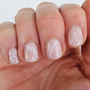 May include: Close-up of a hand with four fingernails painted a light nude color with white leaf designs.