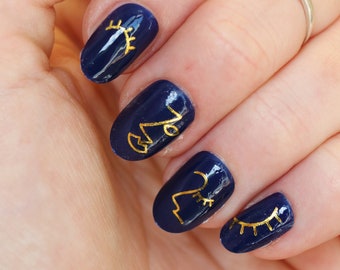May include: Close-up of a hand with navy blue nail polish and gold nail art. The nail art features simple line drawings of eyes and abstract shapes.