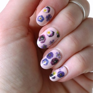 Pode incluir: Close-up de uma mão com esmalte de unha rosa claro e pequenas flores roxas e amarelas pressionadas no esmalte.