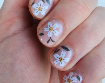 May include: Close-up of a hand with four fingernails painted with a light pink nail polish and decorated with white flowers and green leaves.