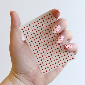 May include: A sheet of nail stickers with a repeating pattern of small red hearts on a white background. The sheet is being held by a hand with pink nail polish and red heart nail art.