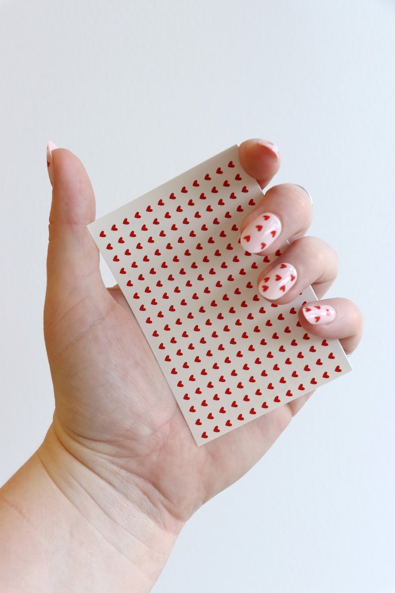 May include: A sheet of nail stickers with a repeating pattern of small red hearts on a white background. The sheet is being held by a hand with pink nail polish and red heart nail art.