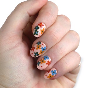 K&ouml;nnte beinhalten: Nahaufnahme einer Hand mit f&uuml;nf Fingern&auml;geln, die mit einer hellrosa Basis und einem floralen Design lackiert sind. Die Blumen sind rot, orange, gelb, blau und gr&uuml;n.