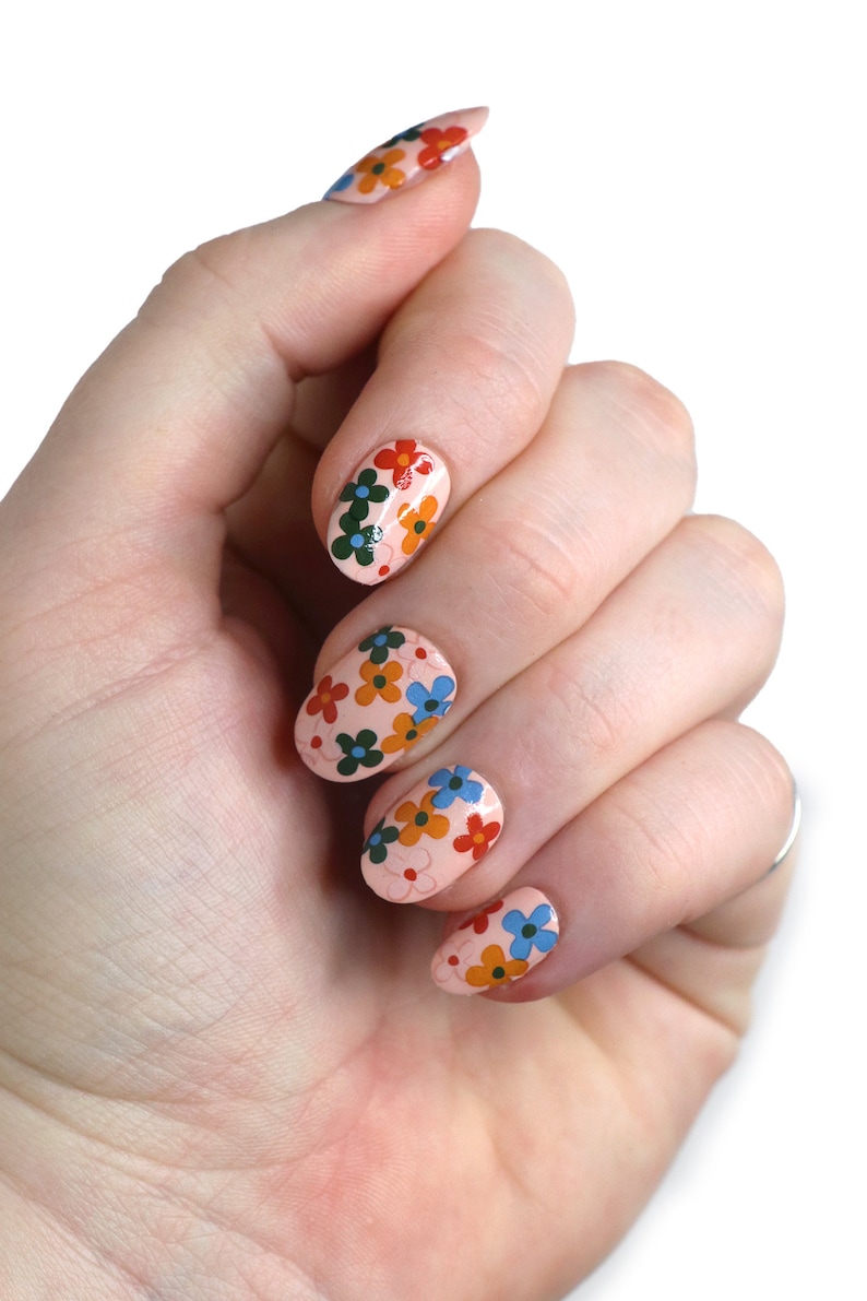 K&ouml;nnte beinhalten: Nahaufnahme einer Hand mit f&uuml;nf Fingern&auml;geln, die mit einer hellrosa Basis und einem floralen Design lackiert sind. Die Blumen sind rot, orange, gelb, blau und gr&uuml;n.