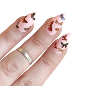 May include: Pink stiletto false nails with butterfly decals. The butterflies are in various shades of brown, orange, and yellow.