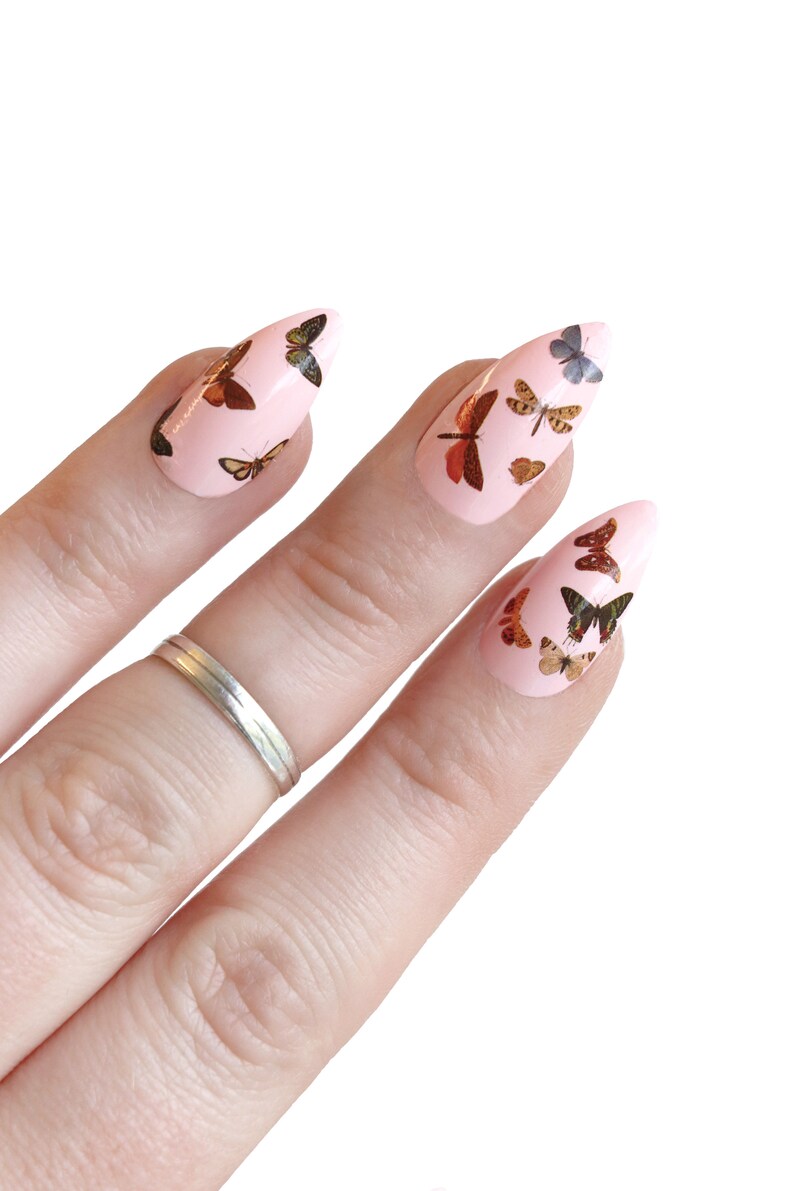 May include: Pink stiletto false nails with butterfly decals. The butterflies are in various shades of brown, orange, and yellow.
