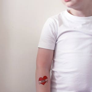 May include: A young person wearing a white t-shirt with a red heart tattoo on their arm. The tattoo has a banner that says "MOM".