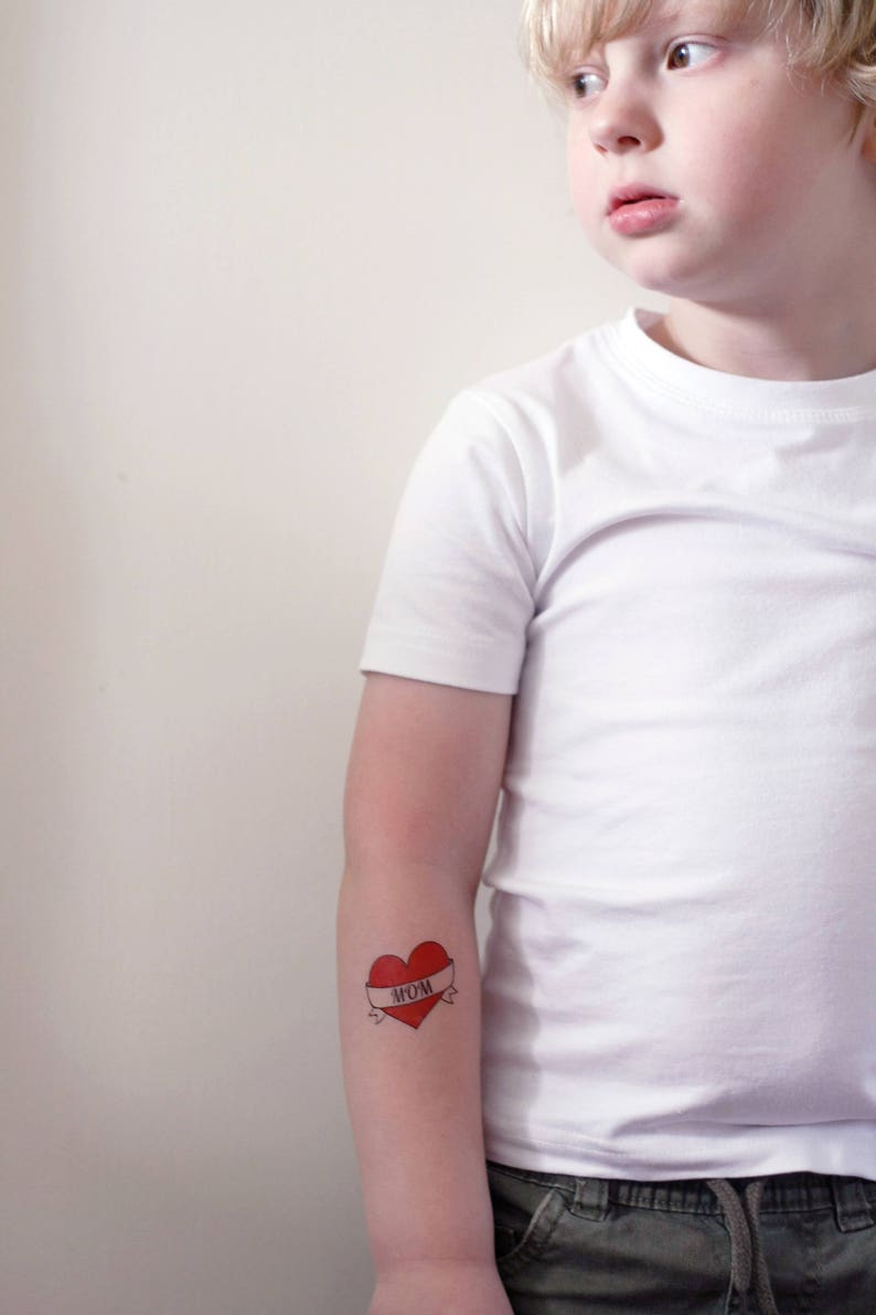 May include: A young person wearing a white t-shirt with a red heart tattoo on their arm. The tattoo has a banner that says "MOM".