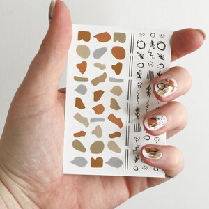 May include: A sheet of nail art stickers with abstract shapes and lines in brown, beige, and gray. The stickers are on a white background.