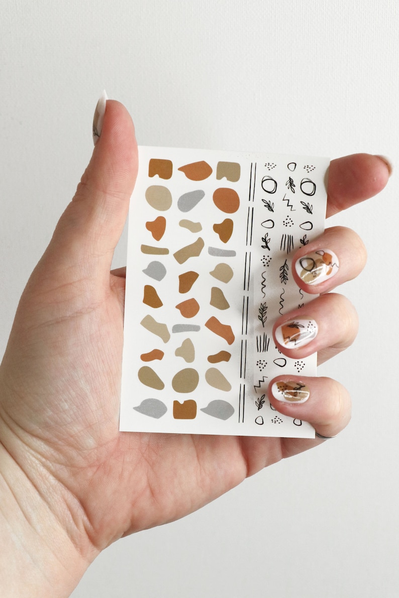 May include: A sheet of nail art stickers with abstract shapes and lines in brown, beige, and gray. The stickers are on a white background.